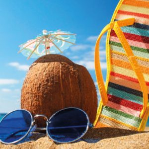 Imagen de una playa donde se ve en primer plano un coco con sombrilla, un par de gafas de sol y parte de una chancla.