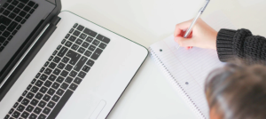 Persona tomando nota y está al lado de un computador portátil.