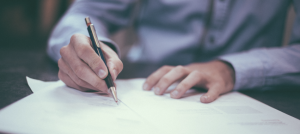 Torse de un hombre sentado a la mesa, escribe sobre hojas blancas.