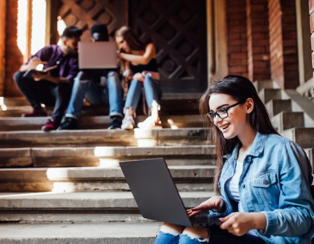 Joven universitaria sentada en unas escalas y sosteniendo un coputador portátil.