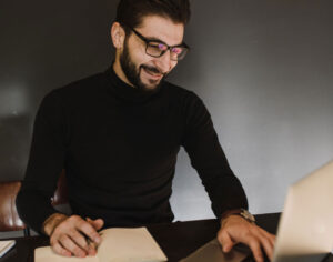 Hombre barbado de gafas y buso negro interactuando con un computador portátil.