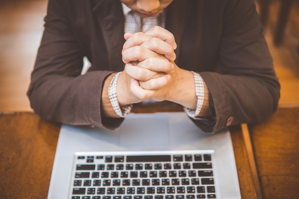 Hombre en actitud de oración en frente de un computador portátil.