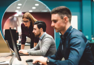 Jóvenes profesionales trabajando frente a computadores en un entorno de oficina, mientras una mujer orienta a uno de ellos.